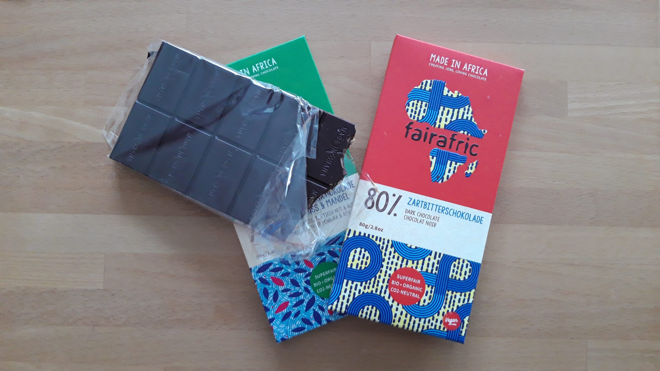 Zwei Tafeln dunkle Fairafric-Schokolade auf Holztisch, eine ausgepackt und angebissen