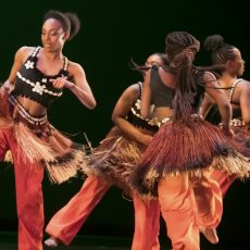 Vier afrikanische Tänzerinnen im Kreis mit orangen Hosen, Baströcken und schwarzen, mit Muscheln besetztem Bustier