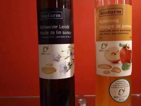 Eine Flasche Schweizer Leinöl und eine Flasche Apfelessig von Biofarm vor roter Wand