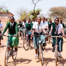 Tansanische MÃ¤dchen in Schuluniform, bestehend aus grÃ¼ndem Jube und weisser Bluse mit grÃ¼ner Krawatte, fahren auf Staubstrasse mit dem Velo zur Schule