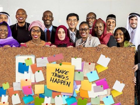Menschen unterschiedlicher Kulturen hinter einer Pinwand, auf der Zettel angebracht sind und in der Mitte die Aufschrift "Make things happen"
