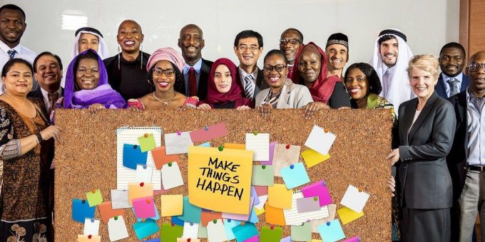 Menschen unterschiedlicher Kulturen hinter einer Pinwand, auf der Zettel angebracht sind und in der Mitte die Aufschrift "Make things happen"
